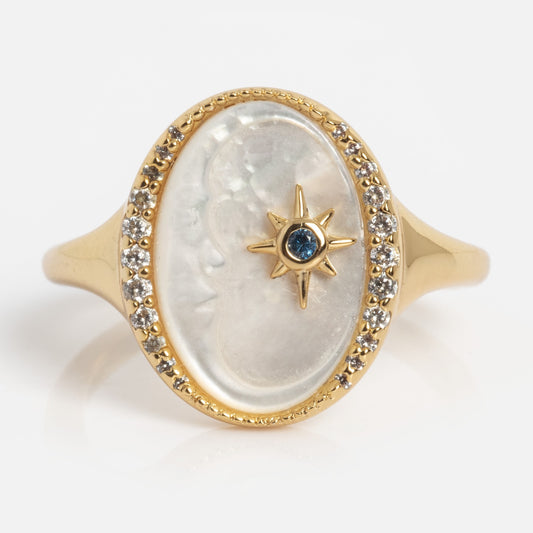 The Carved Moon Mother of Pearl Divination Ring features an oval gold design with a carved moon face on white mother of pearl, encircled by clear gemstones and centered with a gold star holding a blue gem, all set on a polished band.