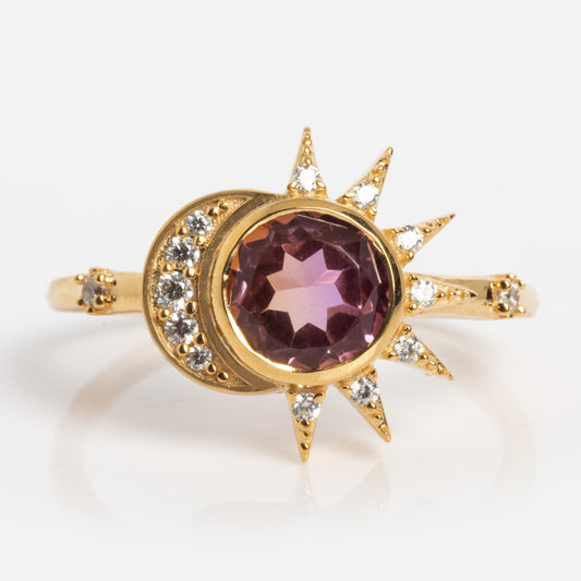 The Divine Duality Sun and Moon Ring is a gold ring with a central round purple gemstone, framed by a crescent moon and sunbeam design accented with small clear stones.
