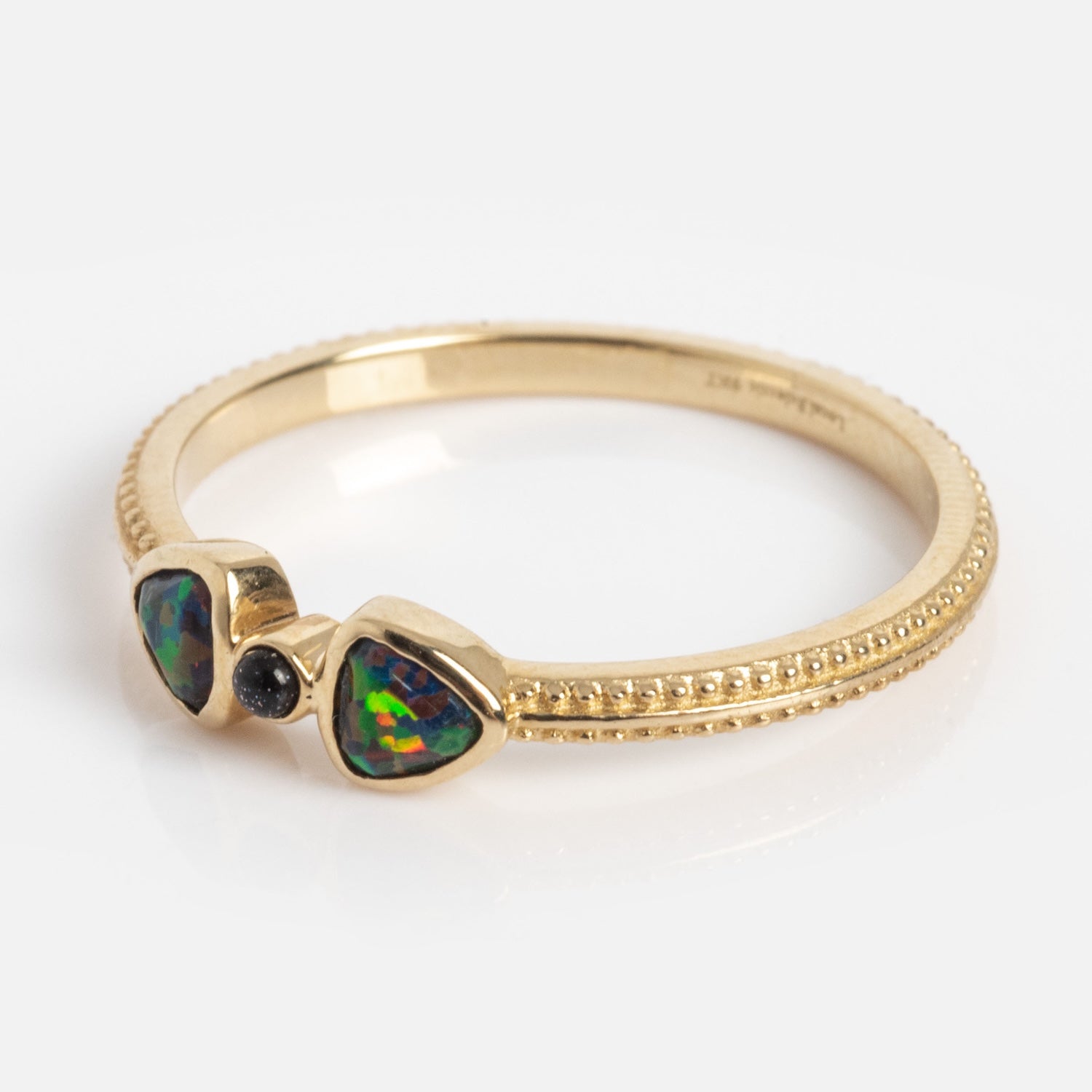 The Solid Gold Black Opal Divine Direction Ring features a textured gold band, two triangular opal stones with green-blue hues, and a small round dark gemstone between them, displayed against a plain white background.