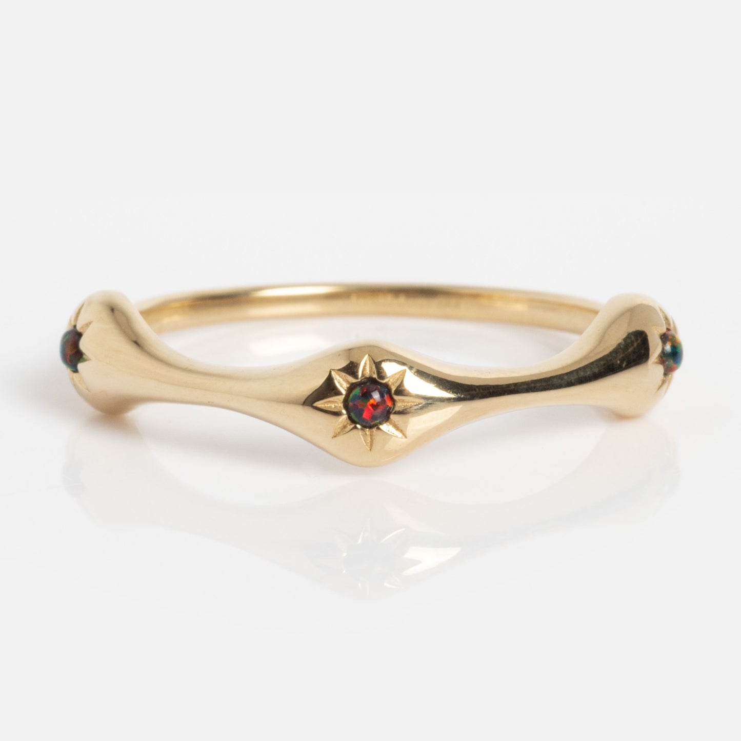 The Solid Gold Black Opal Stars Aligned Ring features a star-shaped setting with a vibrant black opal at its center and small accent stones on each side. The curved gold band is showcased against a white background.