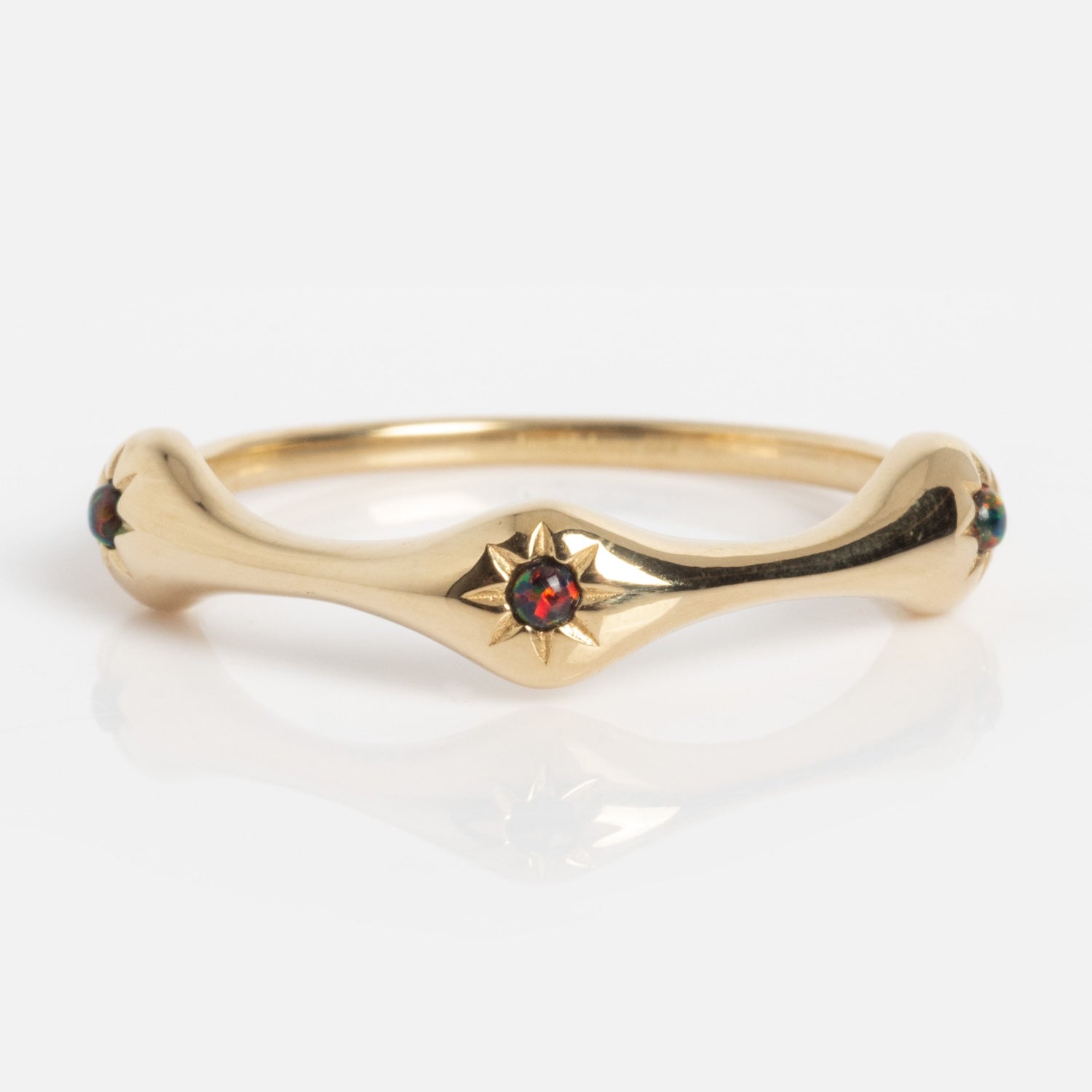 The Solid Gold Black Opal Stars Aligned Ring features a star-shaped setting with a vibrant black opal at its center and small accent stones on each side. The curved gold band is showcased against a white background.