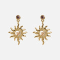 The Summer Solstice Earrings are gold-tone sun-shaped danglers with wavy rays, a round gemstone at the center, and a small gemstone at the top, shown against a plain white background.