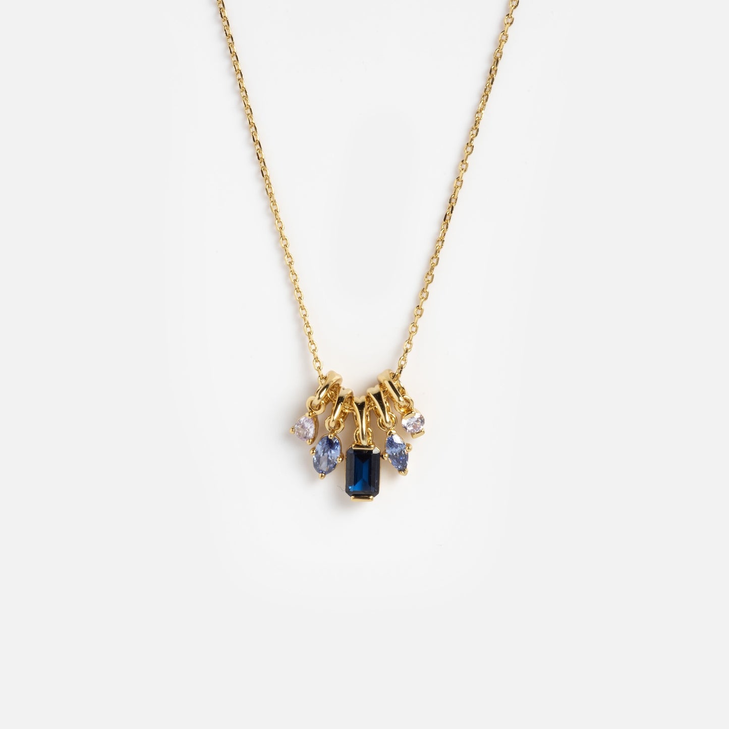 The Shades of You Ombre Birthstone Charm Necklace is a gold chain with five gemstone charms: a central blue rectangular gem, two round clear gems, and two smaller blue stones, all set in gold on a plain white background.
