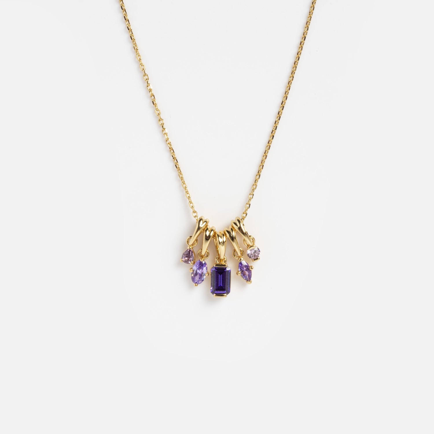 The Shades of You Ombre Birthstone Charm Necklace is a gold chain necklace with five purple gemstone pendants, featuring an emerald-cut center stone and four smaller, variously shaped stones, shown on a white background.