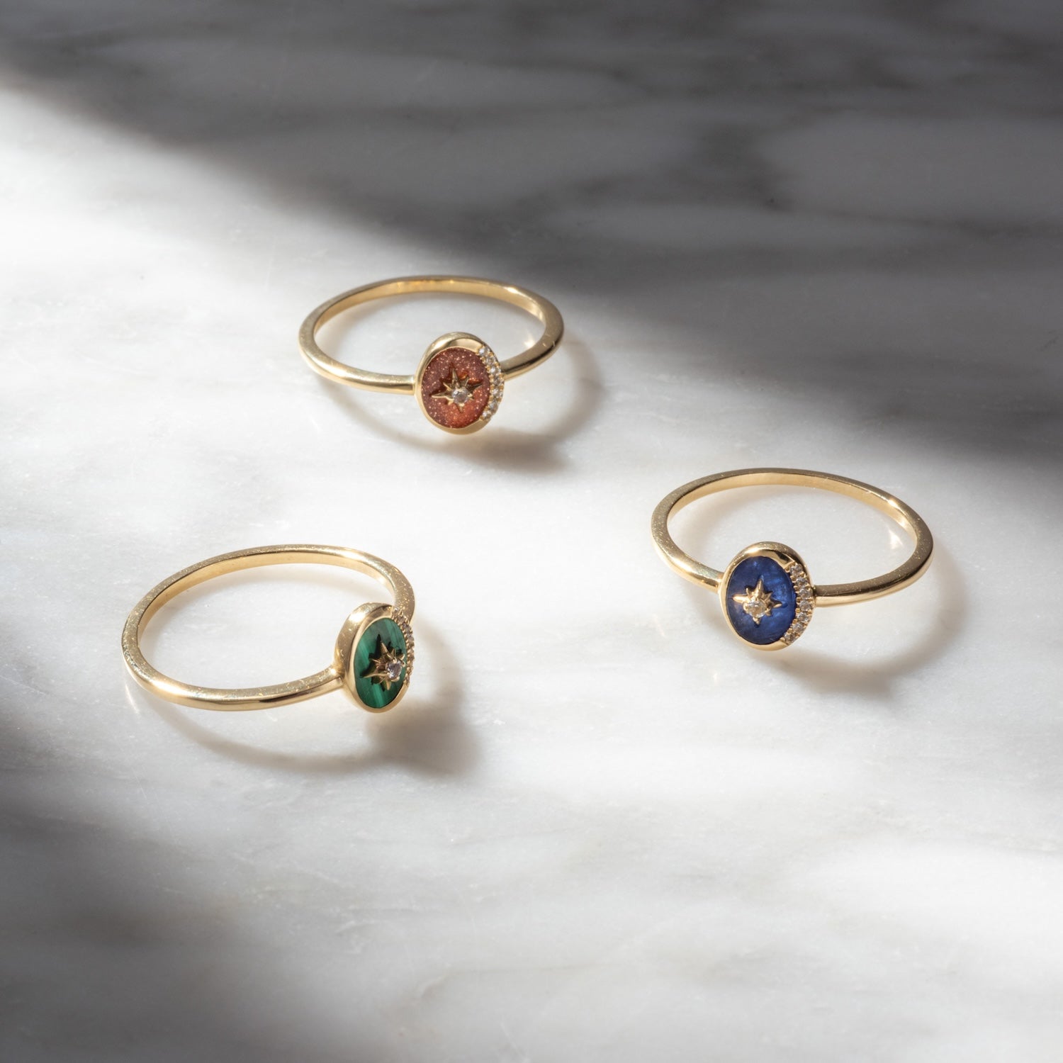 Three Solid Gold Crystal Crescent Moon Rings for Manifestation, with green, red, and blue gemstone settings and a small star detail at the center, are arranged on white marble under soft natural light and shadows.