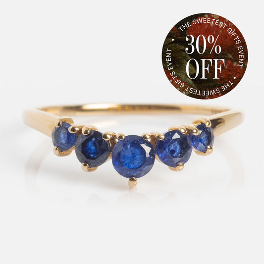The 14k Blue Sapphire Arc Nesting Ring features five round sapphires in a tapered arc design. A circular sticker in the upper right corner says “THE SWEETEST GIFTS EVENT 30% OFF” in white on a dark, multicolored background.