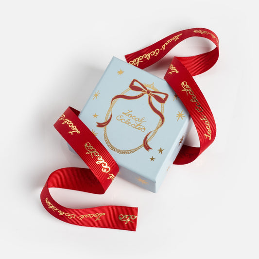 The Limited Edition 2025 Holiday Jewelry Box features pale blue with gold writing, star designs, and a red ribbon adorned with gold text that loops around the box on a white background.