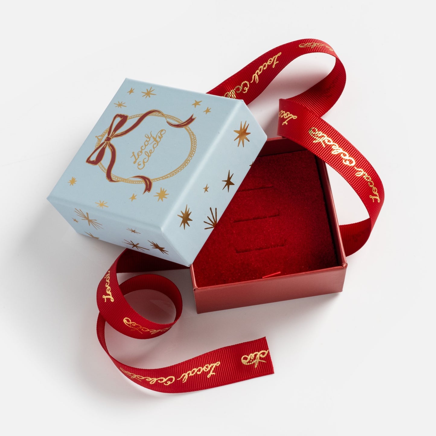 A small, light blue Limited Edition 2025 Holiday Jewelry Box with gold star designs and “lucky charms” on the lid sits open to reveal a red interior. A long red ribbon with gold script is draped around this festive gift box.