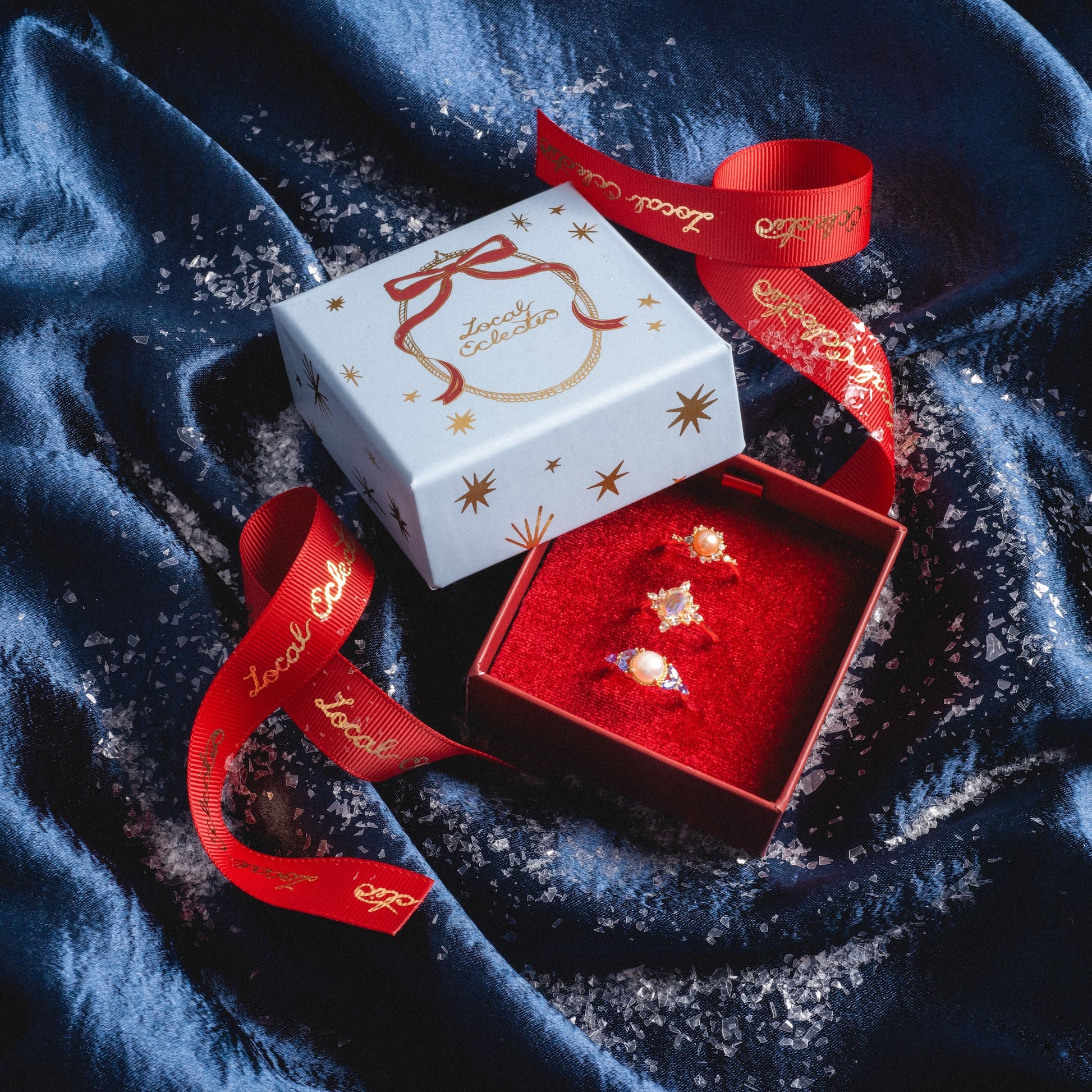 The Limited Edition 2025 Holiday Jewelry Box with star patterns sits open on blue velvet, displaying three gold gemstone rings. Red ribbon with gold lettering and scattered glitter add festive flair to the scene.