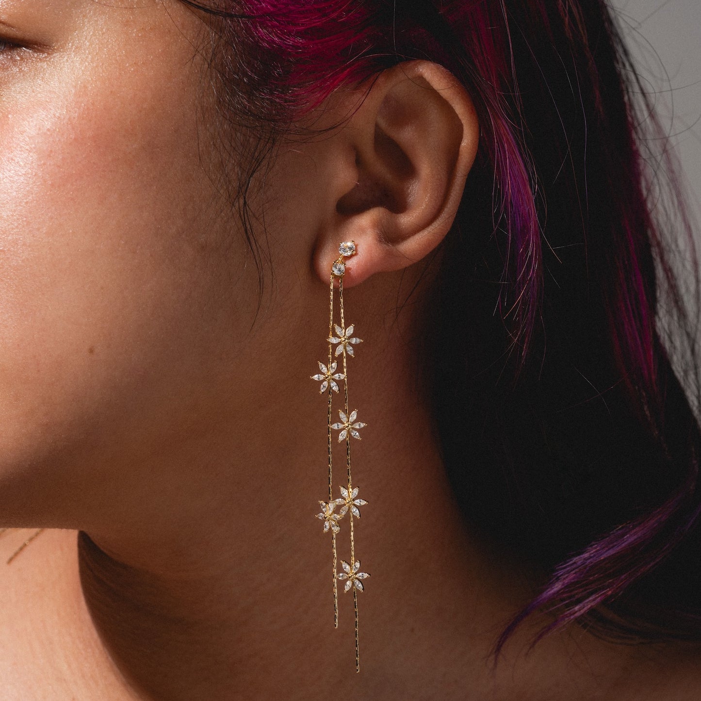 A close-up of a person with magenta hair wearing Whisper Earrings—long, gold, floral-themed danglers adorned with sparkling stones and intricate flower details.
