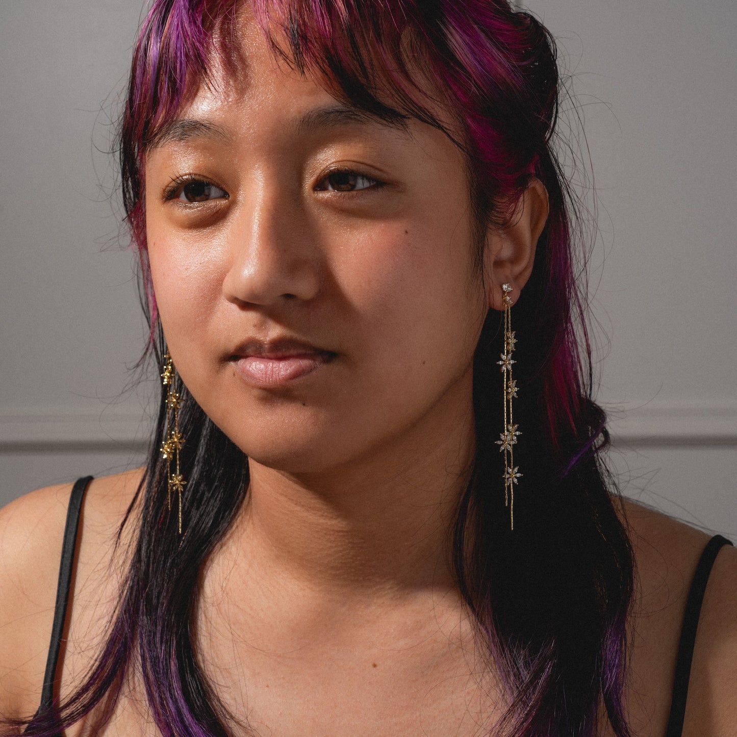 A person with purple-tinted hair and long gold Whisper Earrings looks slightly away from the camera, wearing a black spaghetti strap top against a neutral background.