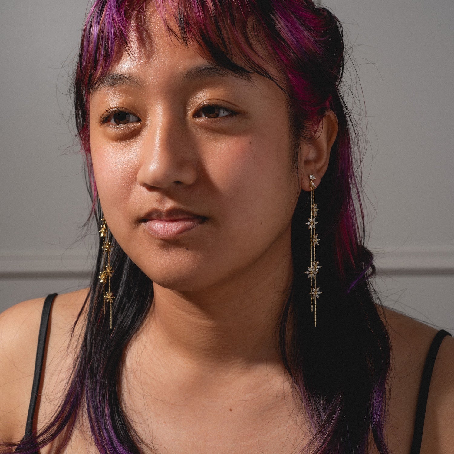 A person with purple-tinted hair and long gold Whisper Earrings looks slightly away from the camera, wearing a black spaghetti strap top against a neutral background.