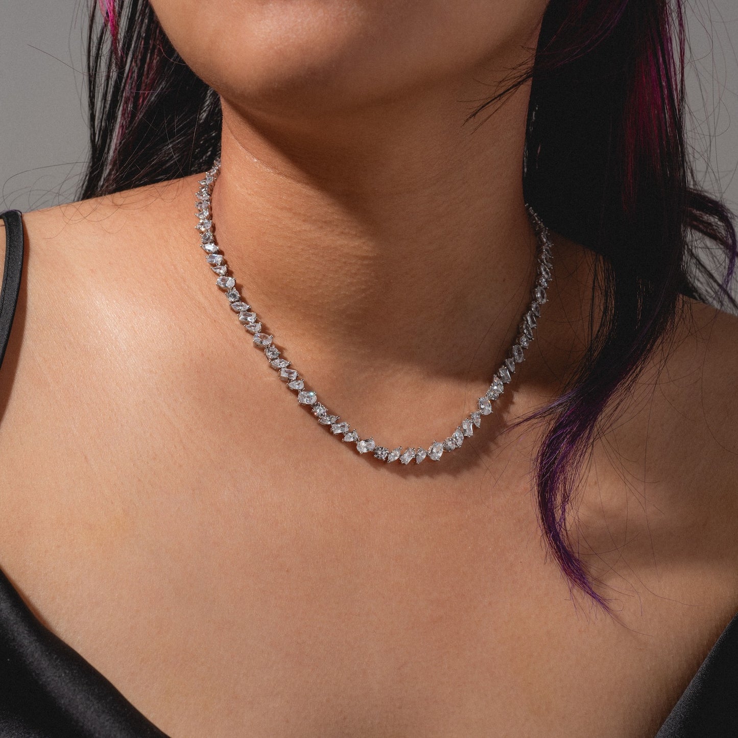 A person with long, dark hair and purple highlights wears a black top and the Pear Diamond Tennis Necklace, which sits closely around the neck and features clear, faceted stones for a sparkly look.