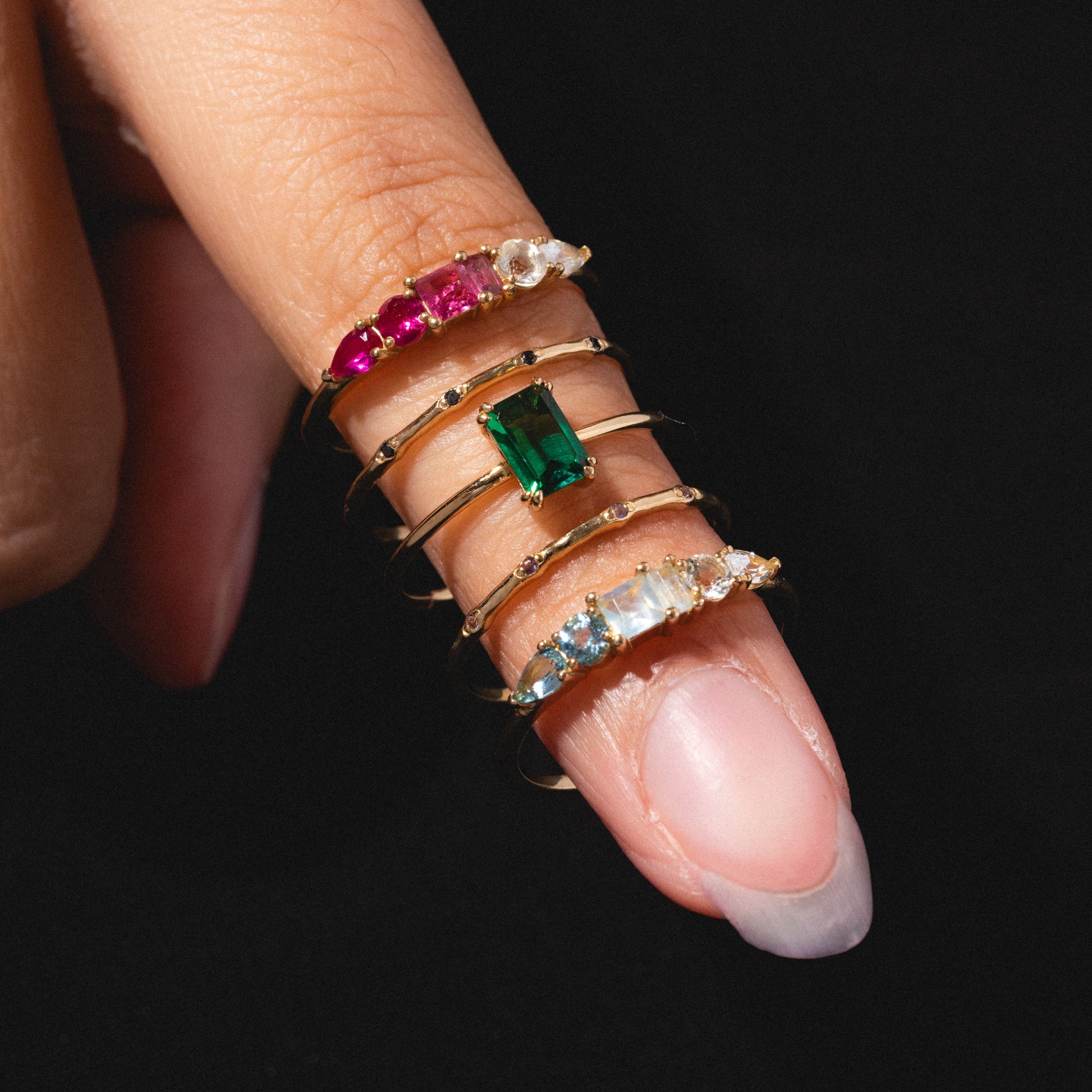 A hand with long, natural nails wears five Solid Gold Dainty Birthstone Stacking Rings—each featuring colorful gemstones in green, red, blue, and clear—against a black background.