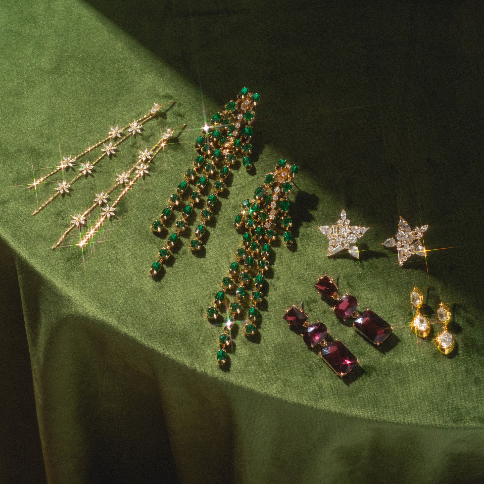 Five pairs of Whisper Earrings are displayed on green velvet, featuring gold star designs, long dangly styles with green gems, purple stones, and round gold and diamond pieces.