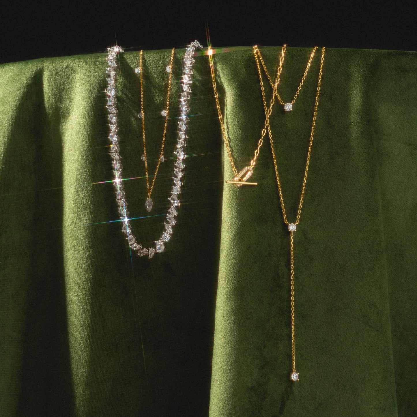 A Pear Diamond Tennis Necklace shines among two delicate gold chains, all elegantly draped over rich green velvet.