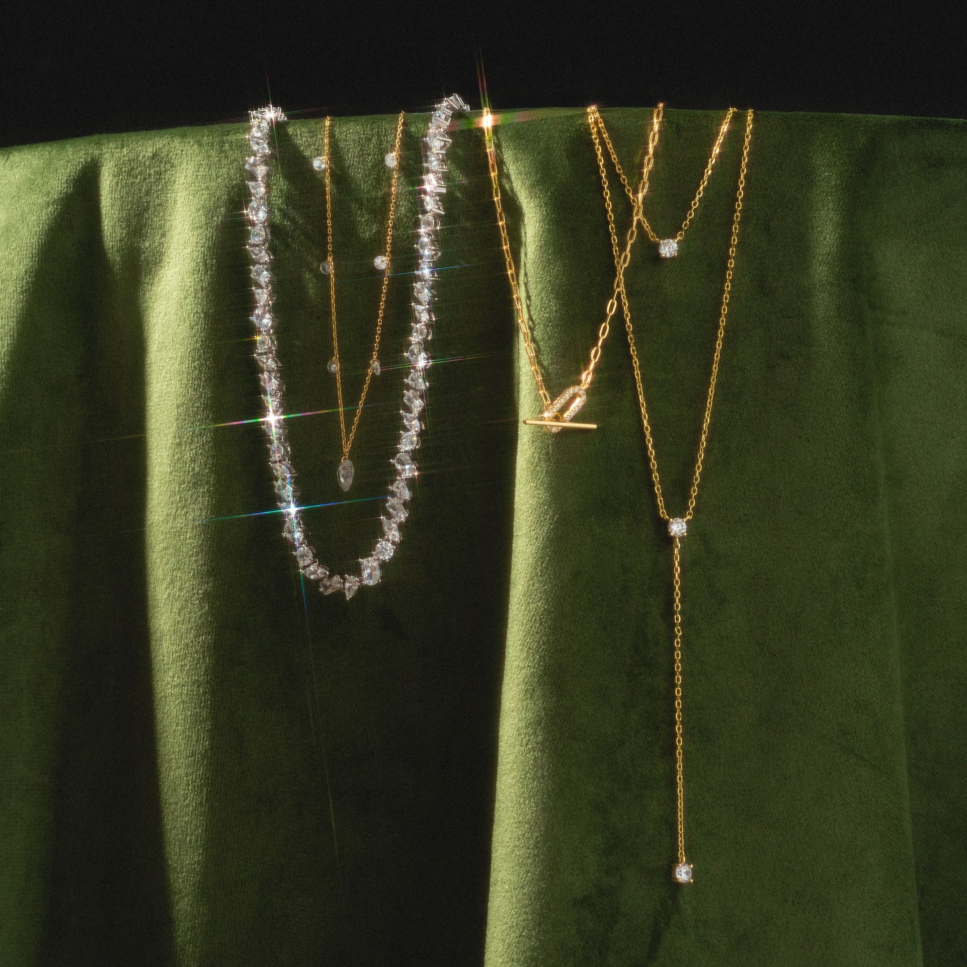 A Pear Diamond Tennis Necklace shines among two delicate gold chains, all elegantly draped over rich green velvet.