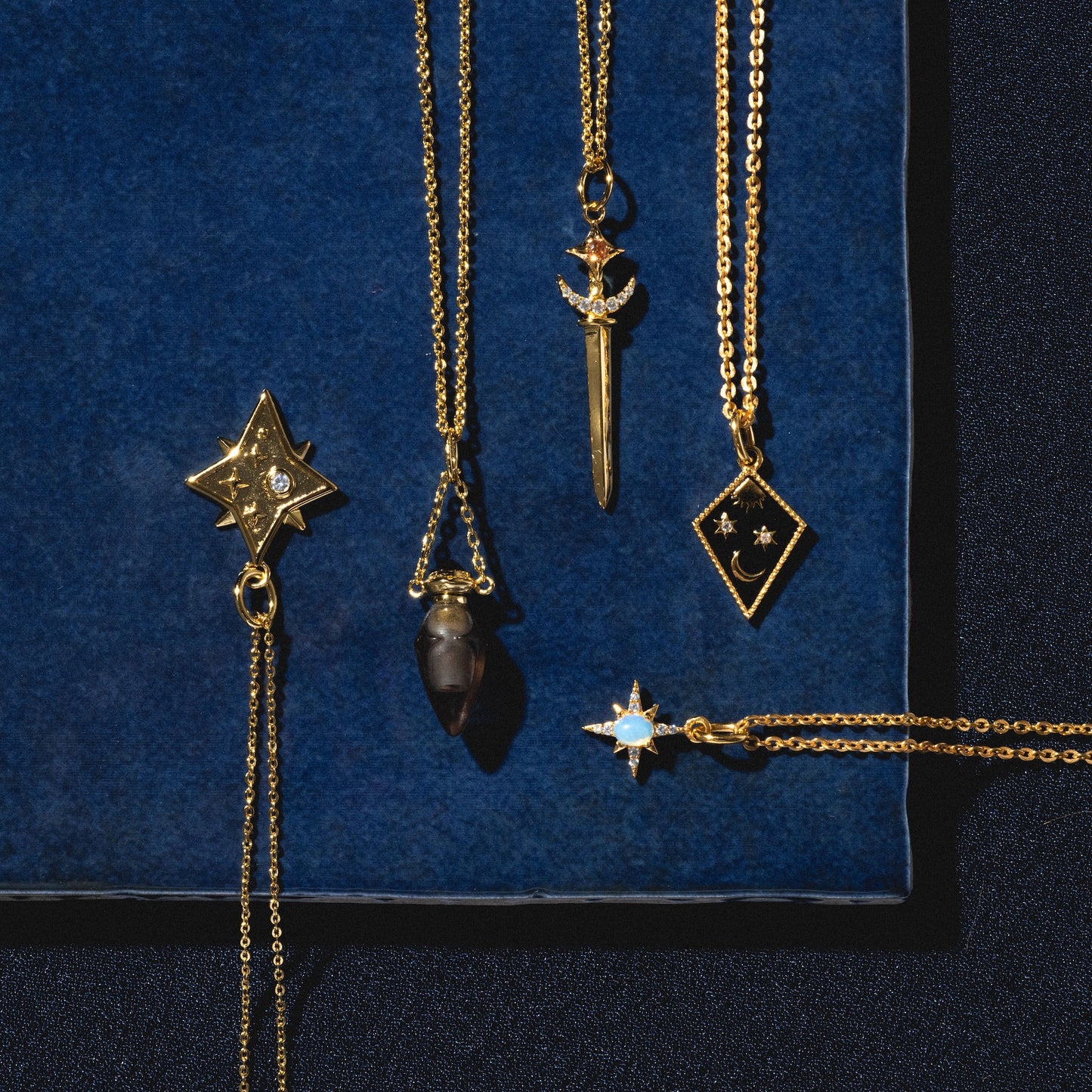 The Sabrina Opal Portal Necklace, a 14k yellow gold plated pendant featuring a simulated opal diamond shape with star accents, is displayed on a dark blue velvet background.