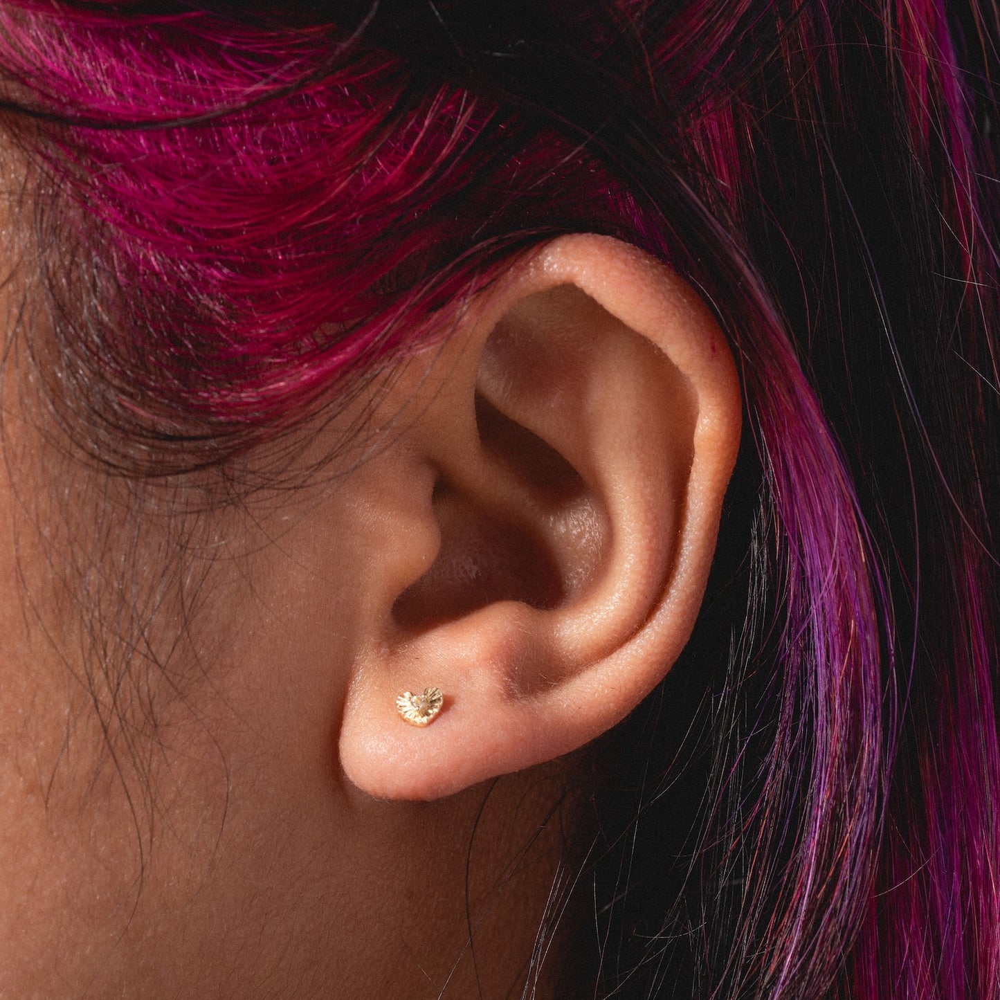 A close-up of a person's ear wearing Solid Gold Heart Flat Back Studs, designed for comfort. The person has dark hair with vibrant magenta highlights.
