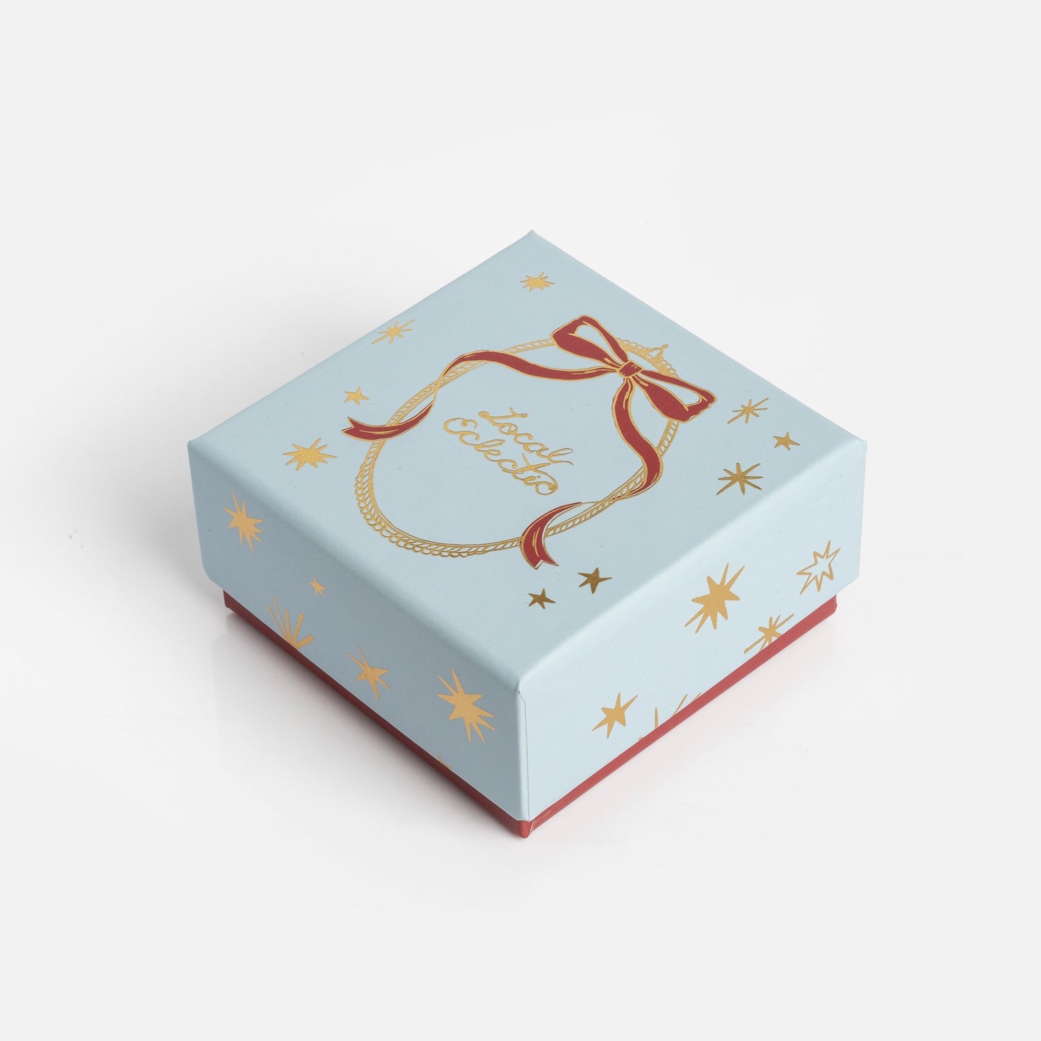 The Limited Edition 2025 Holiday Jewelry Box features a light blue lid with gold stars, a red ribbon illustration, and “Jolly Holiday” in gold script. The box has a red base and is displayed on a white background.