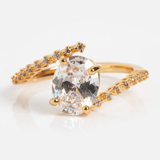 The Tilted CZ Ring is a gold band with a large oval-cut clear CZ centerpiece, split shank, and small round diamonds on each side, displayed against a white background.