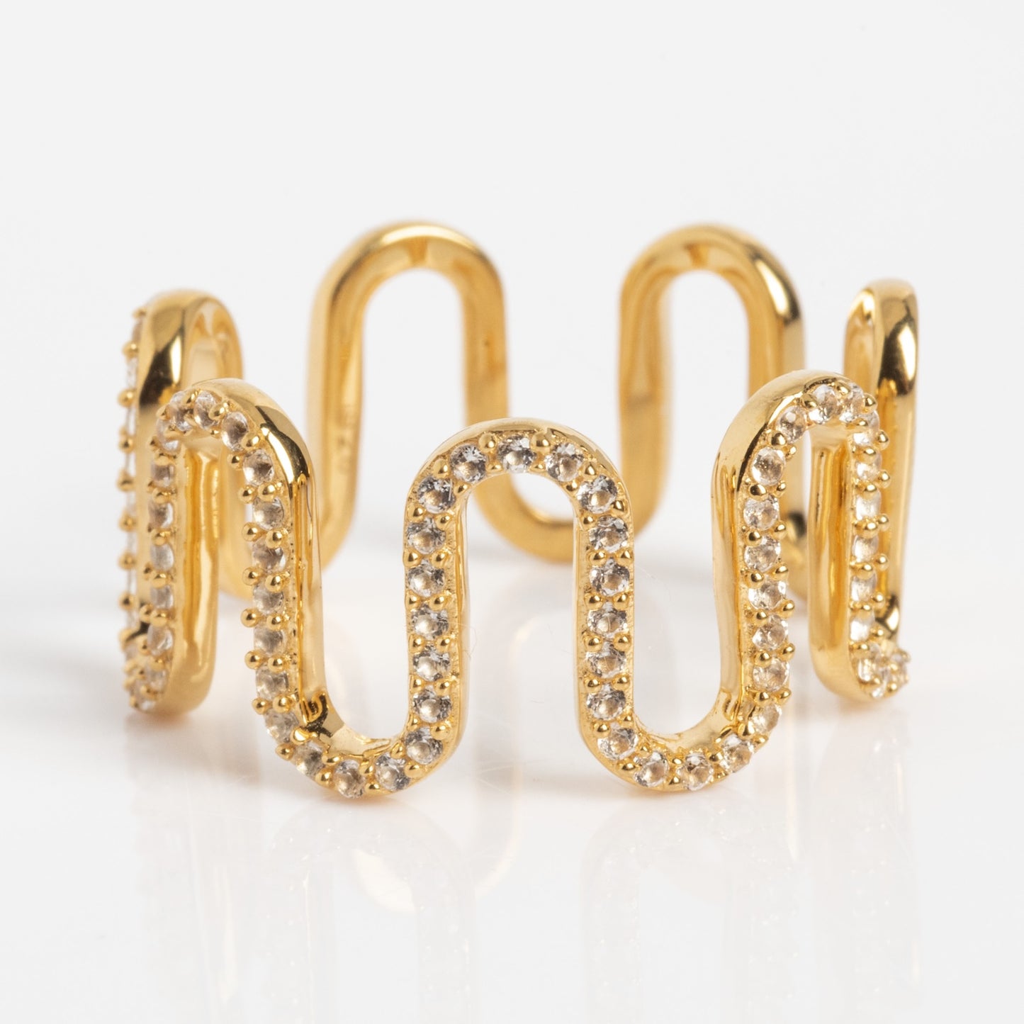 The Curtain Call Cascade Ring is a gold ring featuring a wavy, zigzag design with sparkling white stones along the curves, displayed on a white background.