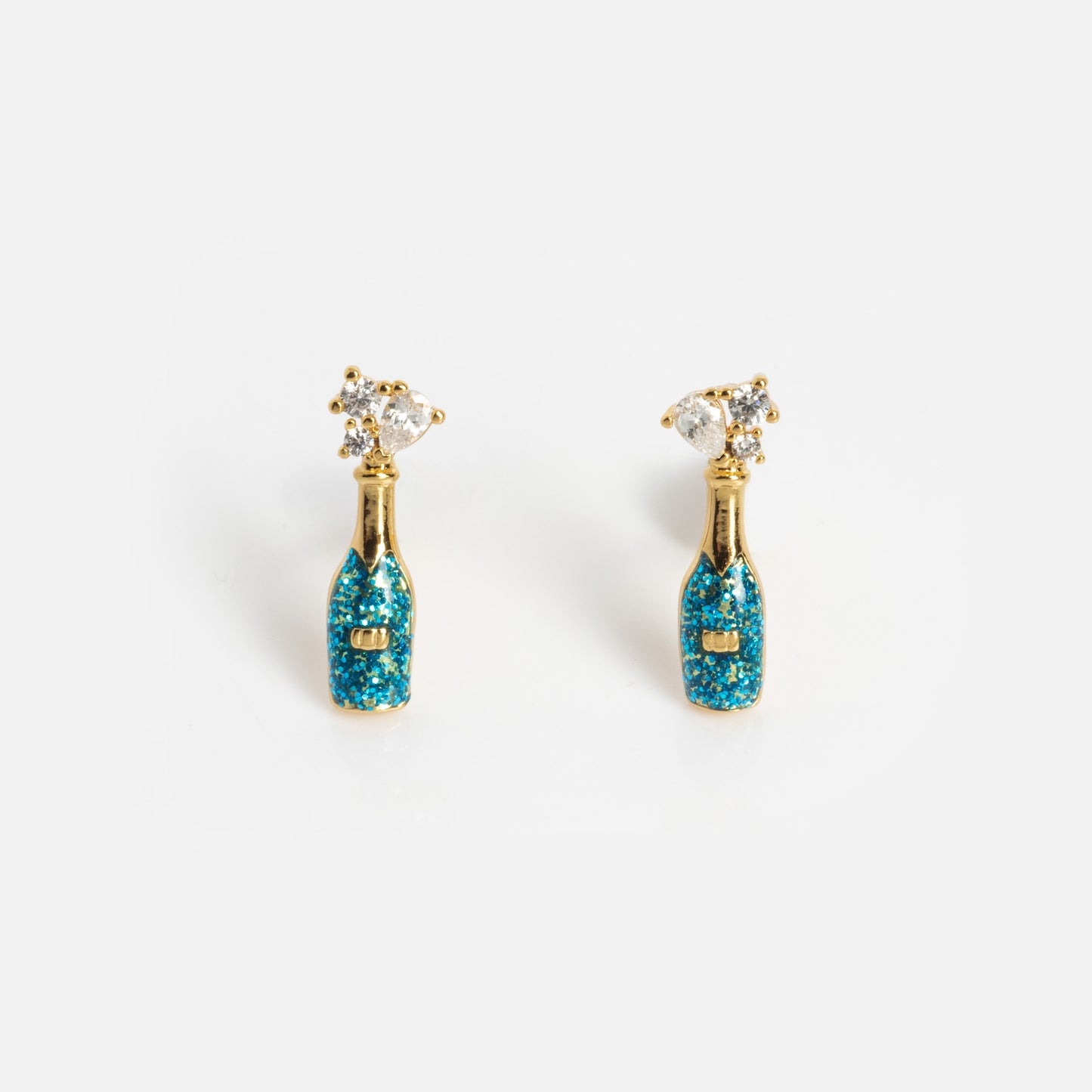 The Champagne Bottle Studs are gold earrings shaped like champagne bottles, featuring blue glittery details and a sparkling crystal popped-cork accent, displayed on a plain white background.