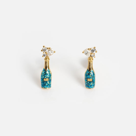 The Champagne Bottle Studs are gold earrings shaped like champagne bottles, featuring blue glittery details and a sparkling crystal popped-cork accent, displayed on a plain white background.