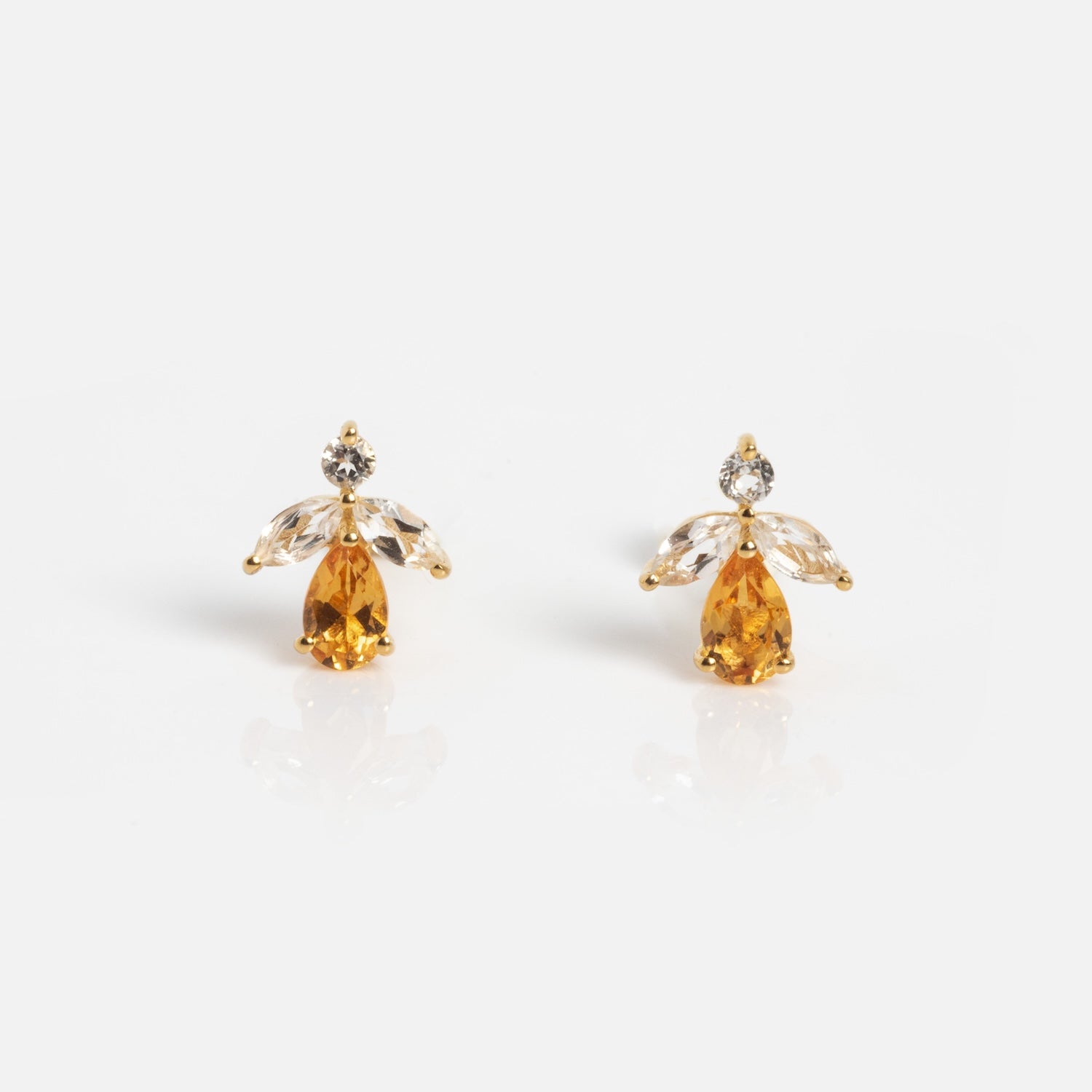 The Jazz Age Citrine Studs are gold earrings shaped like angels, with a yellow teardrop gem for the body, two clear marquise stones as wings, and a round clear gem as the head, all set against a white background.