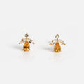 The Jazz Age Citrine Studs are gold earrings shaped like angels, with a yellow teardrop gem for the body, two clear marquise stones as wings, and a round clear gem as the head, all set against a white background.