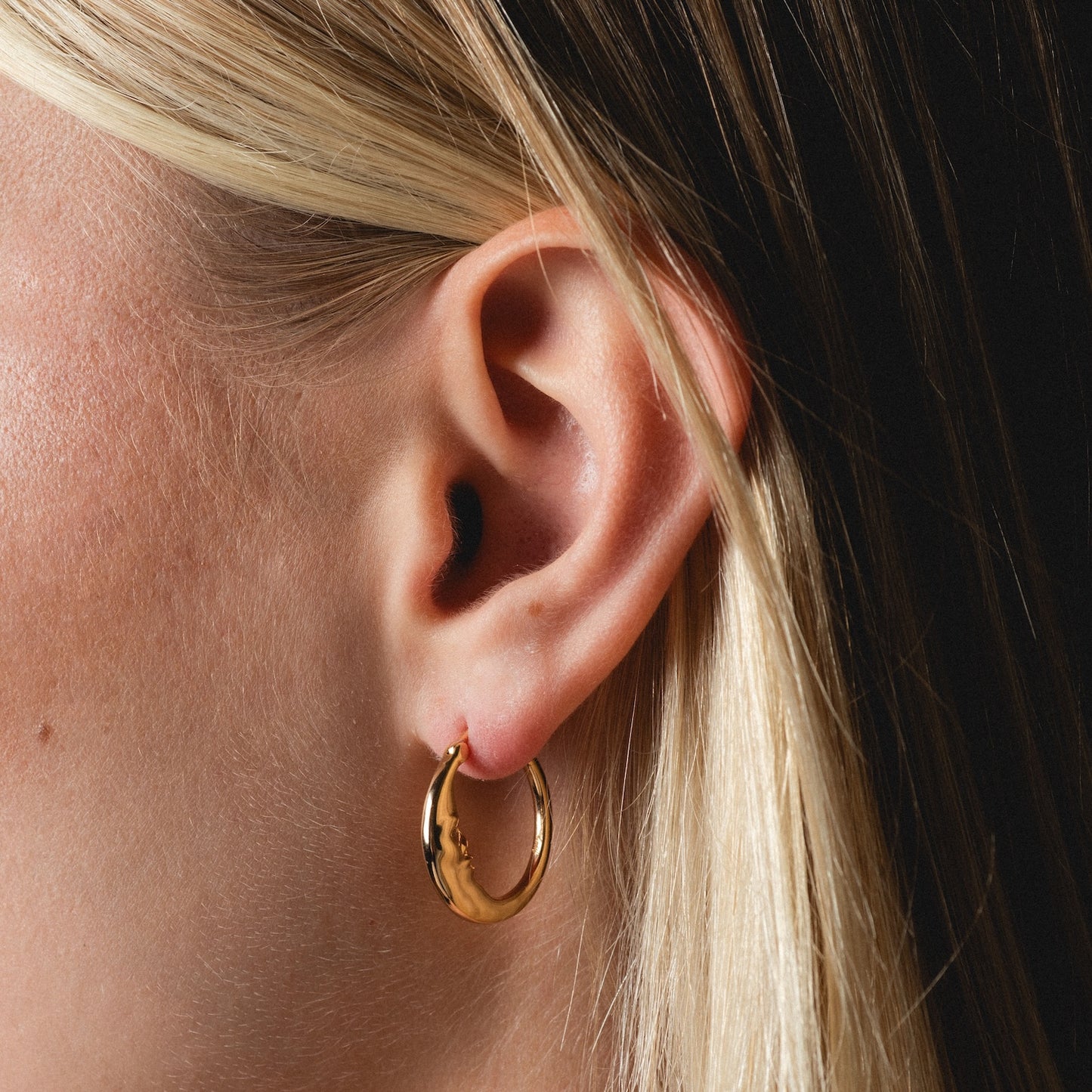 Close-up of a person with straight blonde hair wearing shiny Petite Luna Hoops gold earrings.