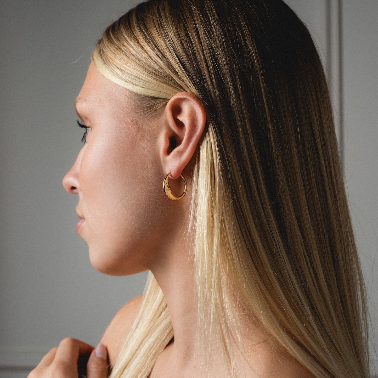 A woman with straight, long blonde hair is shown in profile wearing the Petite Luna Hoops. The softly lit, neutral background highlights the gold earrings.