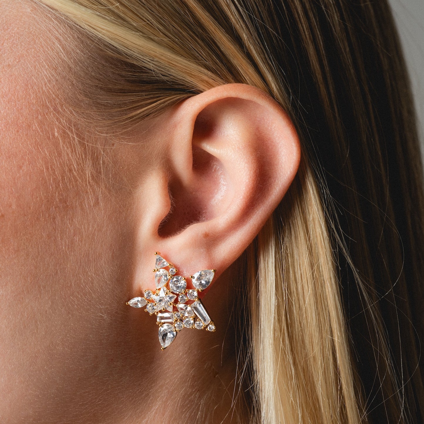 Close-up of a person's ear wearing Debut Star Stud Earrings, large and star-shaped with sparkling gemstones. Their straight blonde hair is tucked behind the ear.
