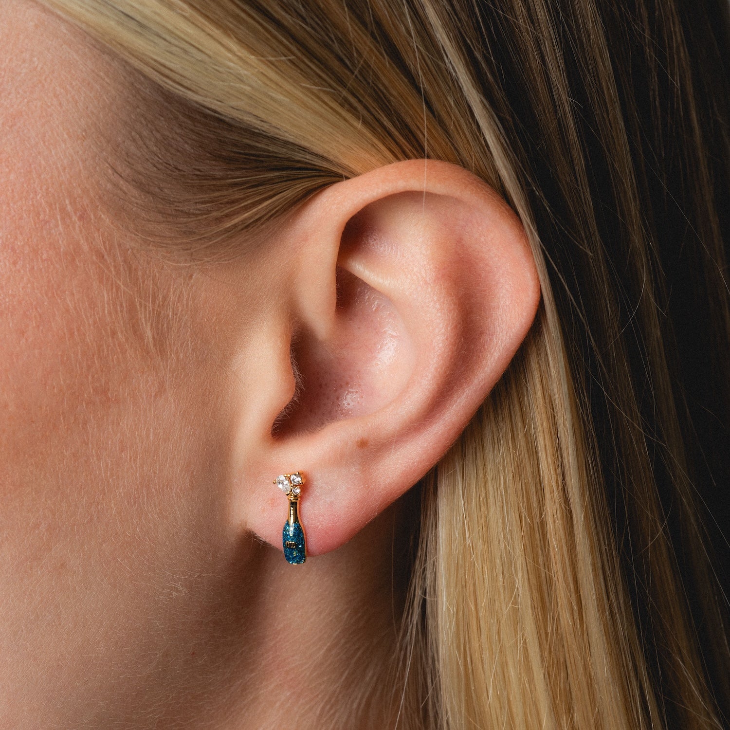 Close-up of a woman's ear with straight blonde hair, wearing Champagne Bottle Studs—small gold earrings topped with a clear stone and a dangling blue teardrop-shaped gem.