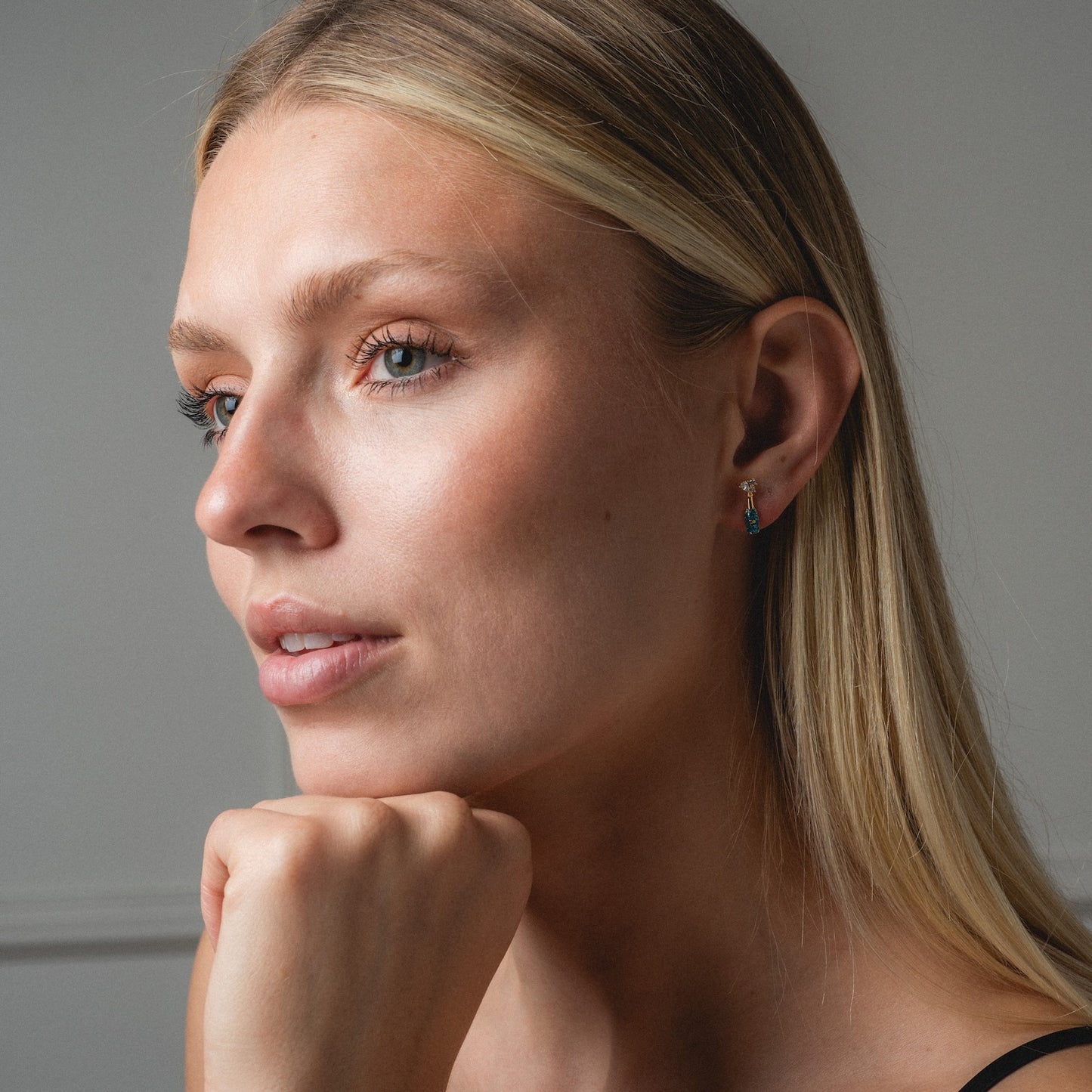 A woman with long blonde hair and natural makeup gazes thoughtfully to the side, resting her chin on her hand. She wears Champagne Bottle Studs with a black strap top against a neutral background.