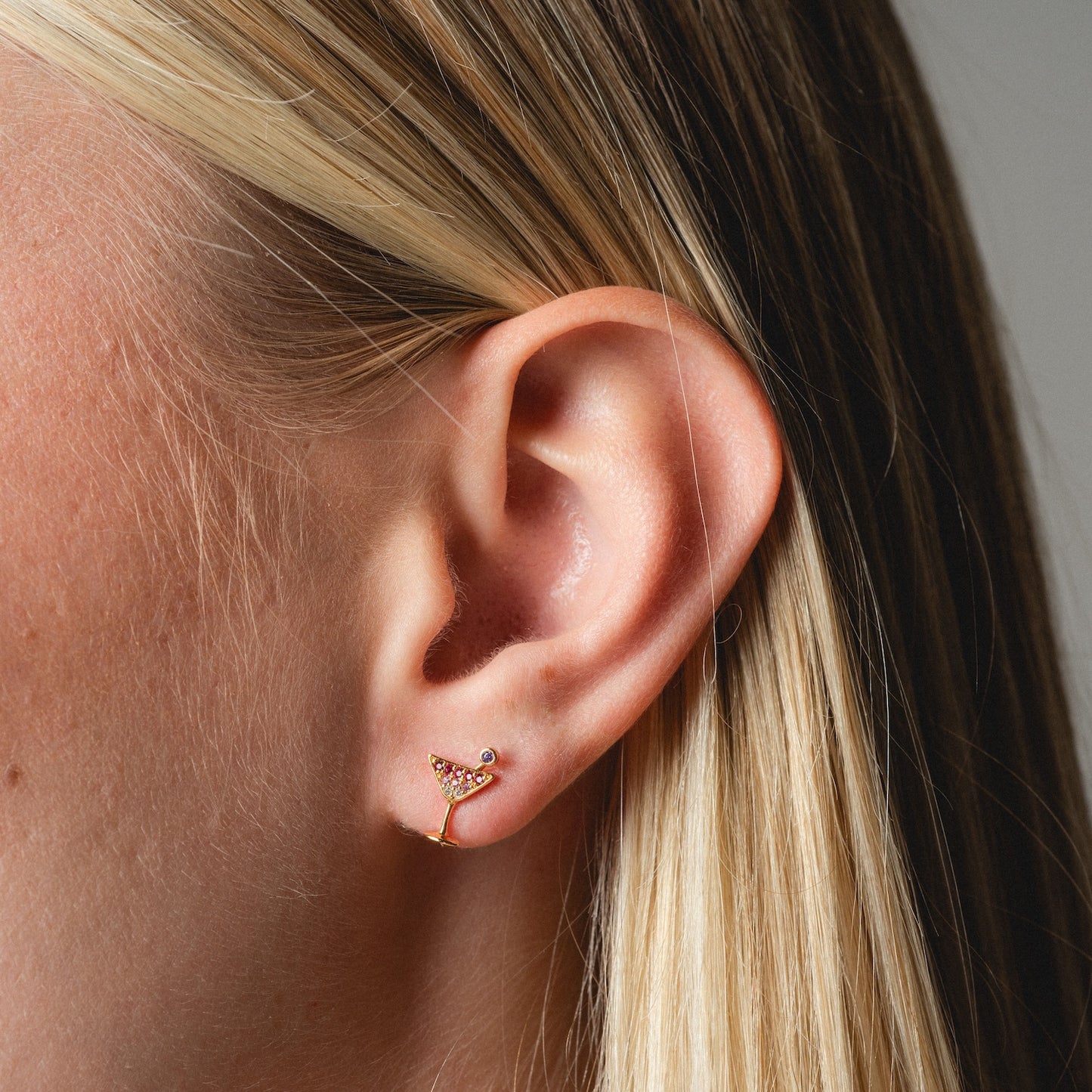 Close-up of a person's ear with straight blonde hair, wearing Martini Glass Studs—small gold earrings shaped like martini glasses with gem details—and a tiny round stud above.