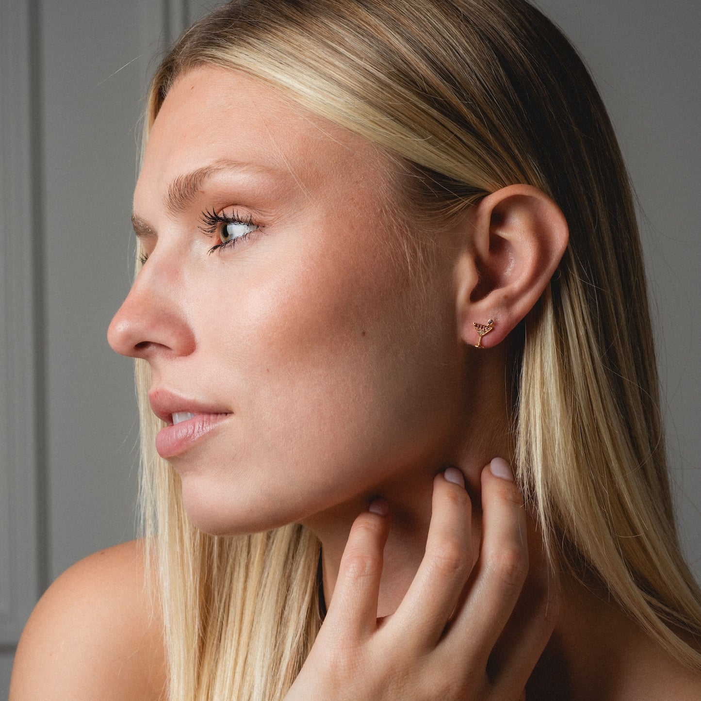 A woman with long blonde hair gazes left, displaying delicate Martini Glass Studs. Her hand rests on her neck and she wears a calm, neutral expression against a softly blurred background.