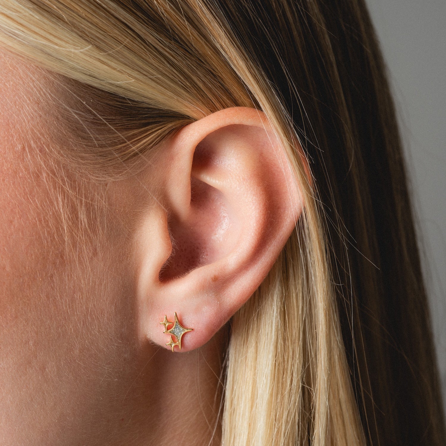 Close-up of a person's ear with straight blonde hair, wearing Twinkling Star Studs—small gold star-shaped earrings. The neutral background is softly blurred.
