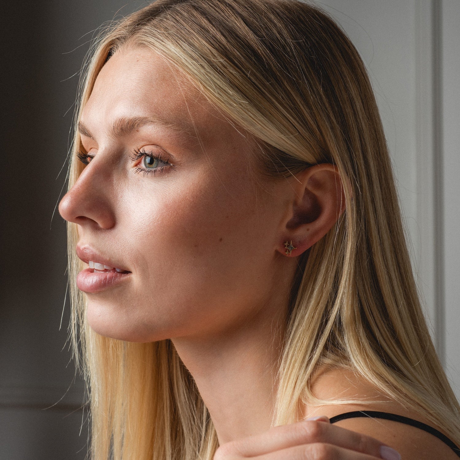 A woman with long blonde hair gazes left in soft natural light, wearing Twinkling Star Studs and a black top, her expression calm and thoughtful.