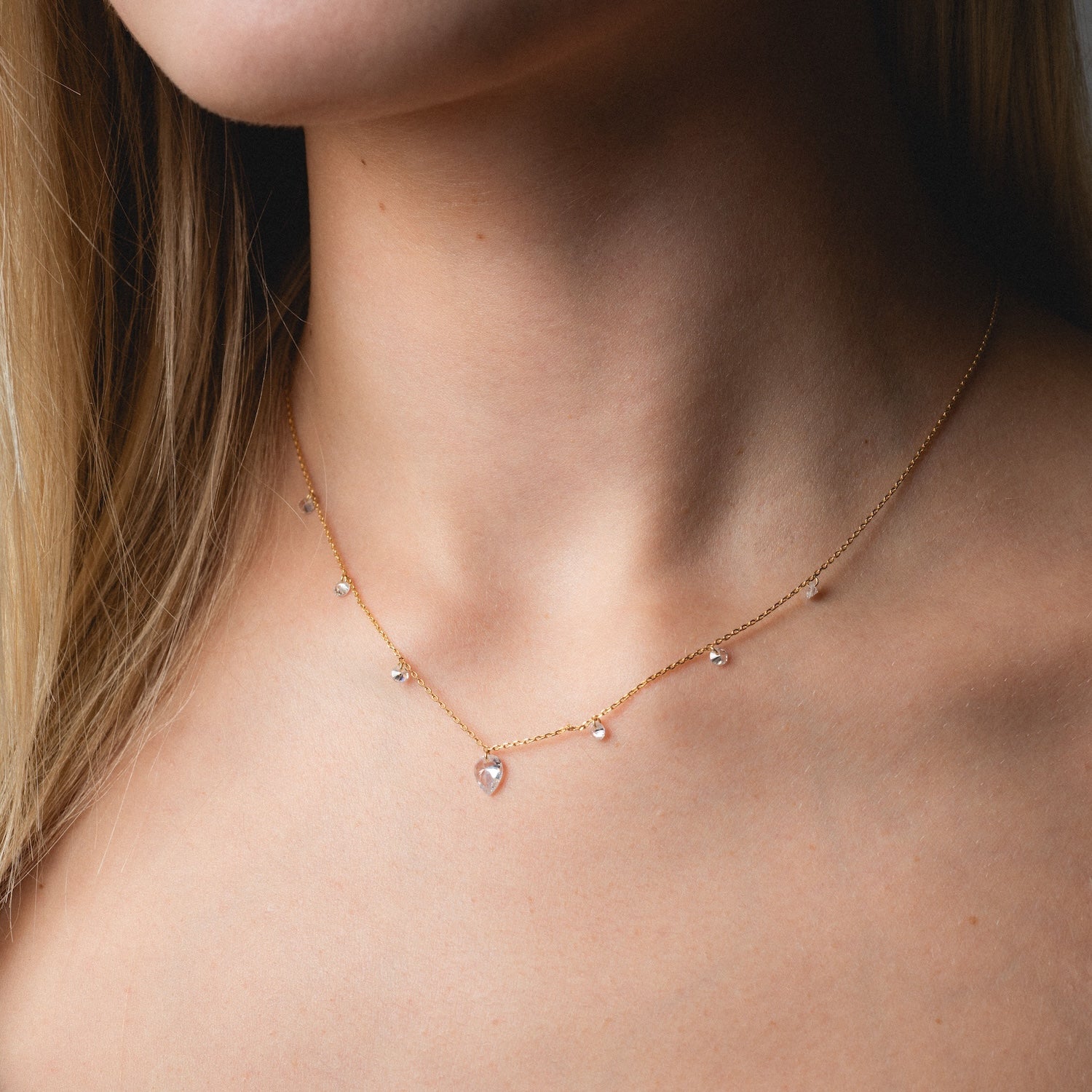 A close-up shows a woman with blonde, straight hair and bare shoulders wearing the Floating CZ Station Necklace—a delicate gold chain featuring small cubic zirconia pendants and a teardrop-shaped central charm.