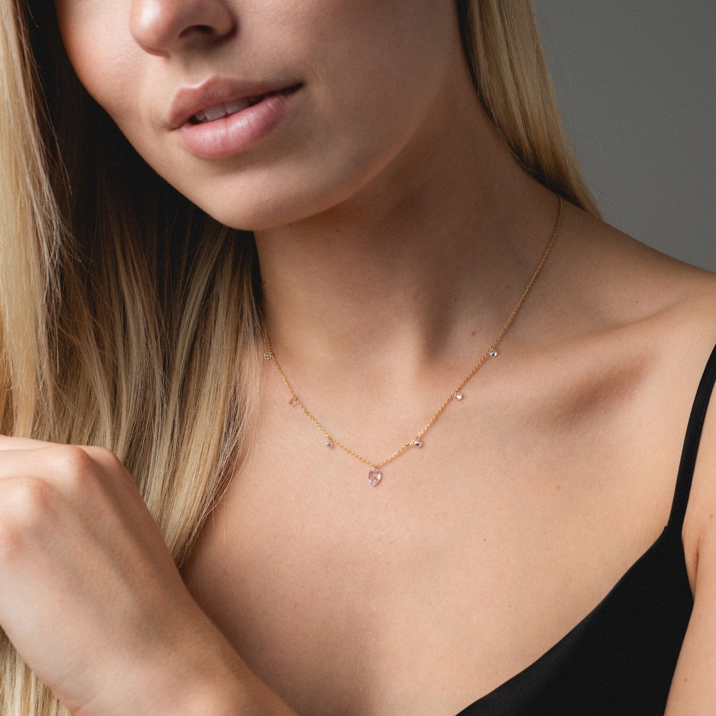 A woman with long blonde hair wears the Floating CZ Station Necklace, featuring small gemstones and a heart pendant. She is dressed in a black spaghetti strap top, shown from the shoulders up against a neutral background.
