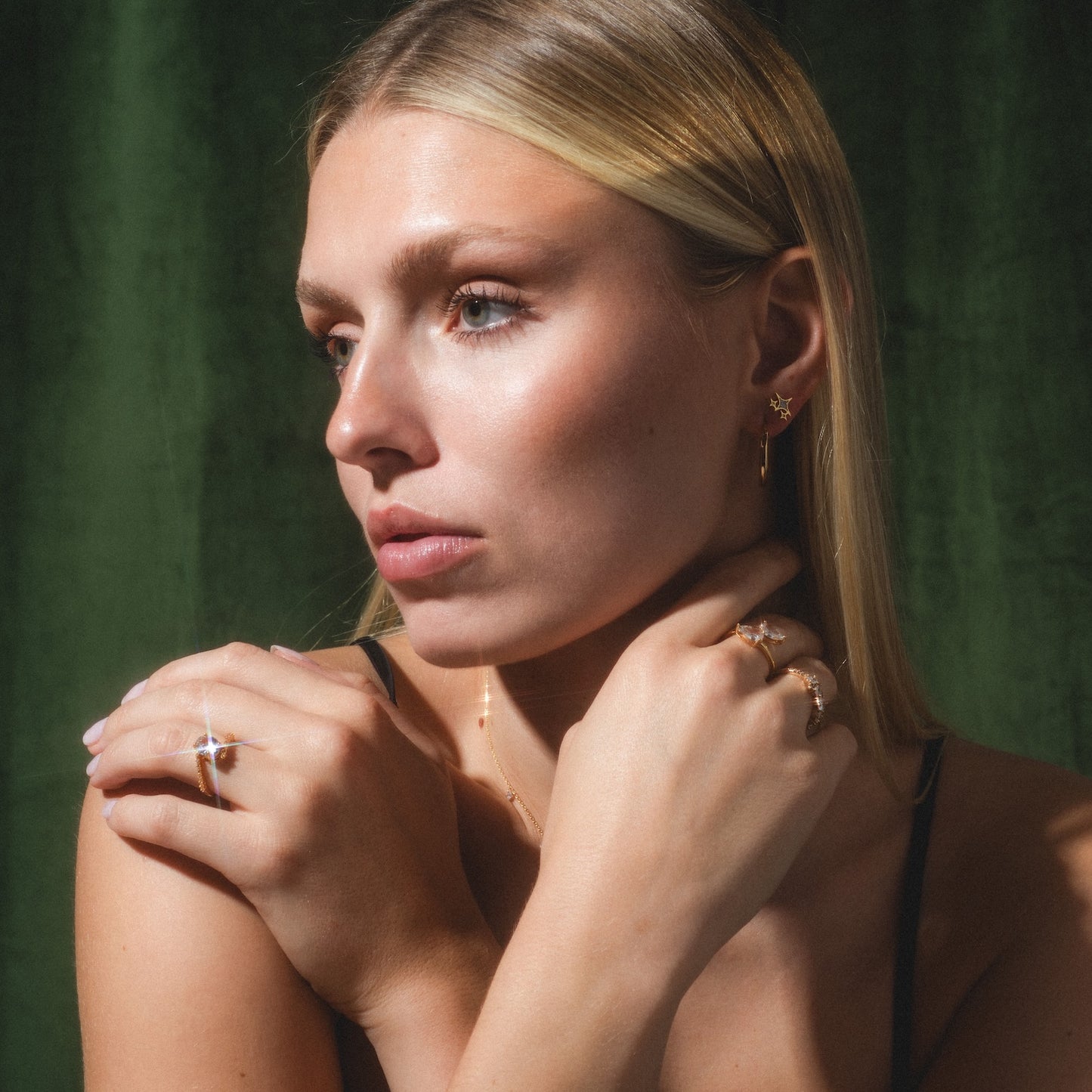 A woman with straight blonde hair poses against a dark green background, wearing Twinkling Star Studs earrings, gold rings, and a necklace. She gazes to the side with her hands gently crossed near her neck.