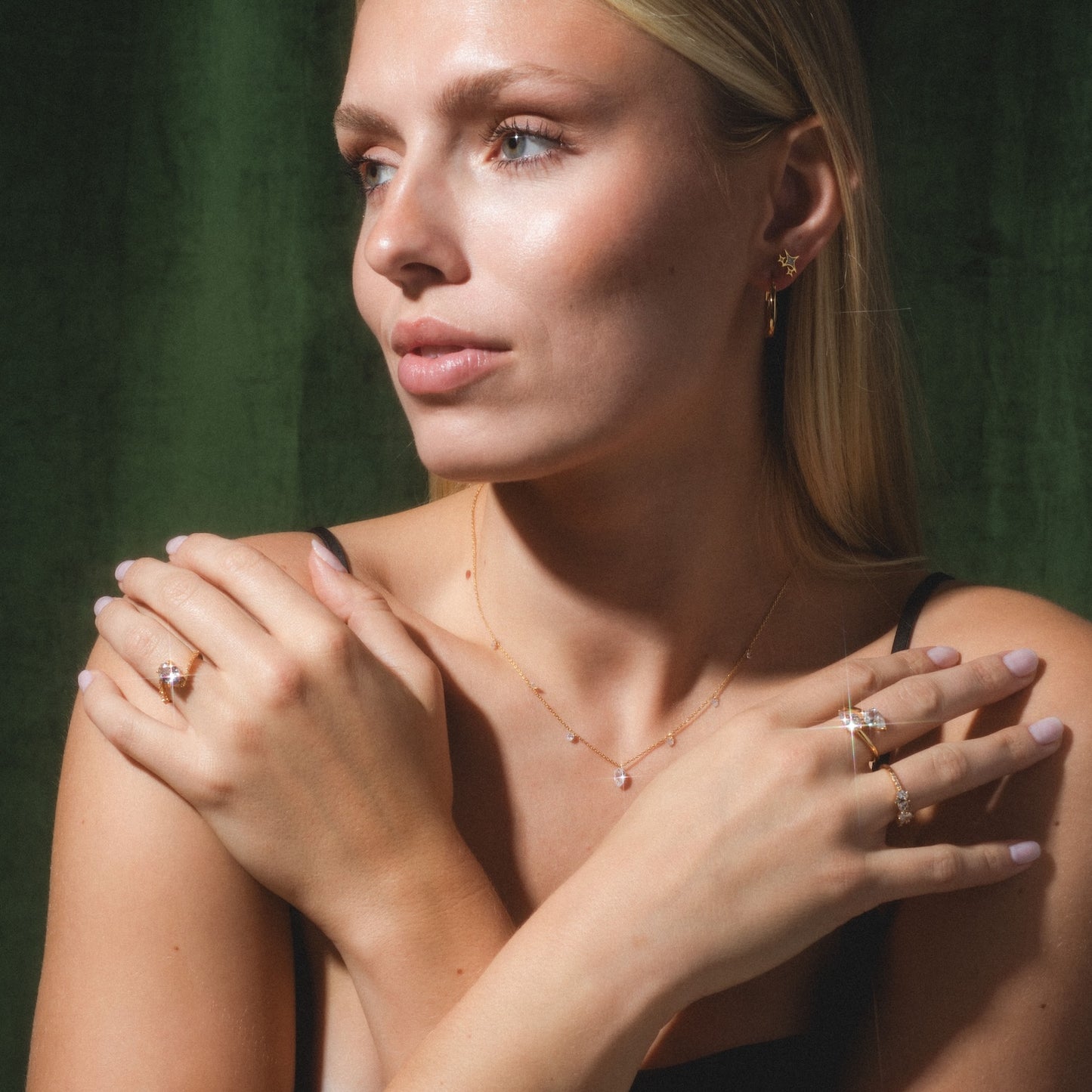 A blonde woman with radiant skin poses before a dark green backdrop in a black top, showcasing the Floating CZ Station Necklace along with gold rings, earrings, and a delicate bracelet as she crosses her arms.