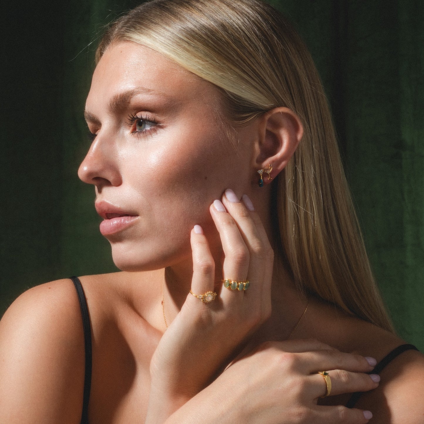 A woman with long blonde hair poses against a dark green background, wearing the Serena Ring along with gold earrings and a necklace. She rests her manicured hand on her face, showcasing the elegant Serena Ring and a calm expression.