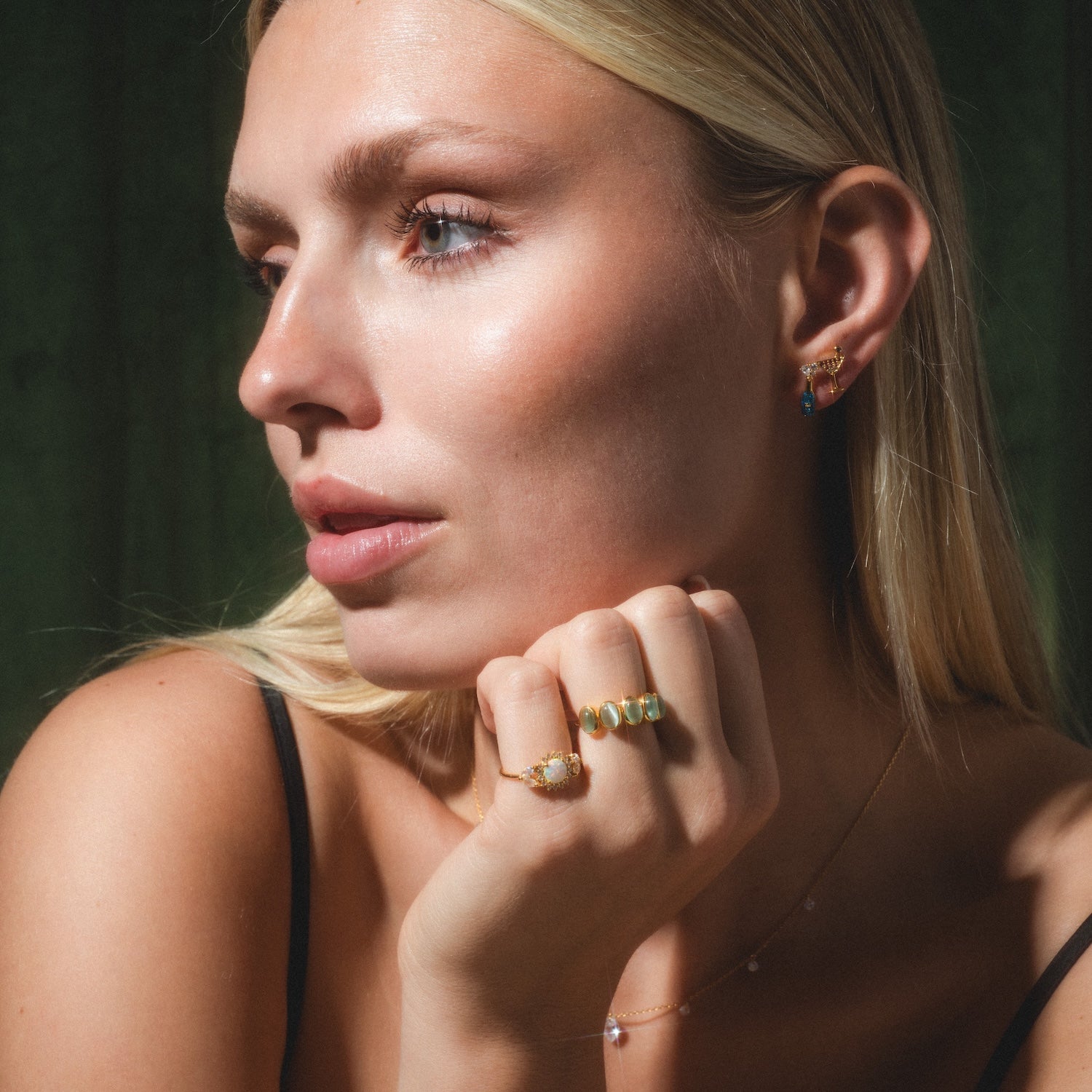 A woman with long blonde hair gazes to the side, wearing Martini Glass Studs, rings, and a delicate necklace. She has glowing skin, nude makeup, and gently touches her face against a dark green background.