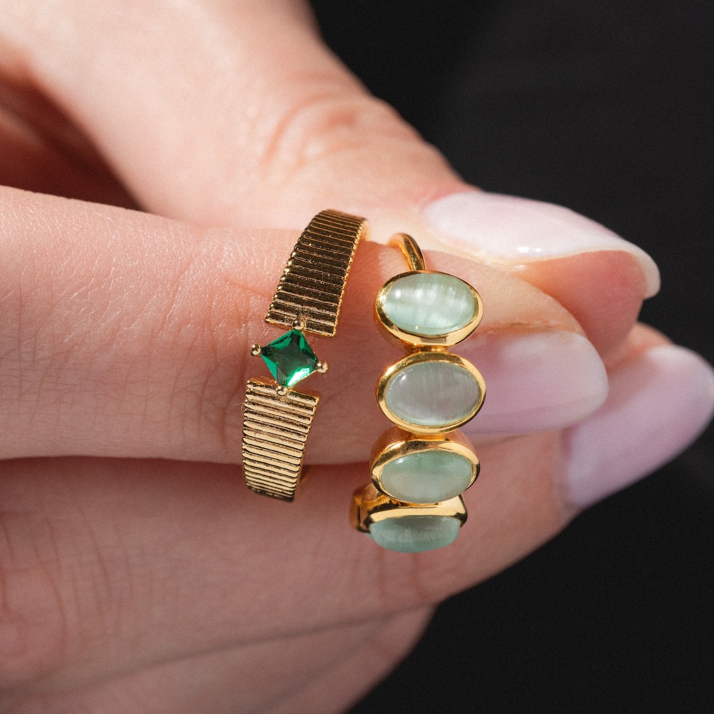 A hand with pale pink polished nails holds two gold rings, including the Sky Blue Cats Eye Flow Ring, which features three oval, sky blue cat’s eye stones in a row.