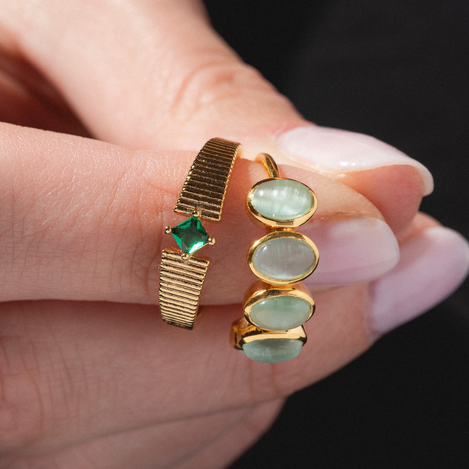 A hand with pale pink polished nails holds two gold rings, including the Sky Blue Cats Eye Flow Ring, which features three oval, sky blue cat’s eye stones in a row.
