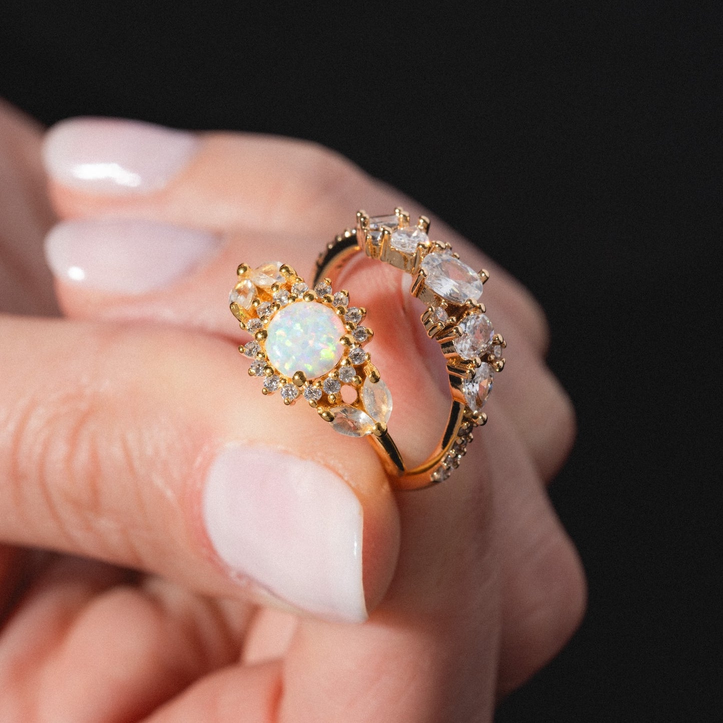 A hand with manicured nails holds the Statement Crystal Cluster Ring, featuring a gold band with a sparkling opal center, surrounded by diamonds and clear gemstones. The background is dark and out of focus.