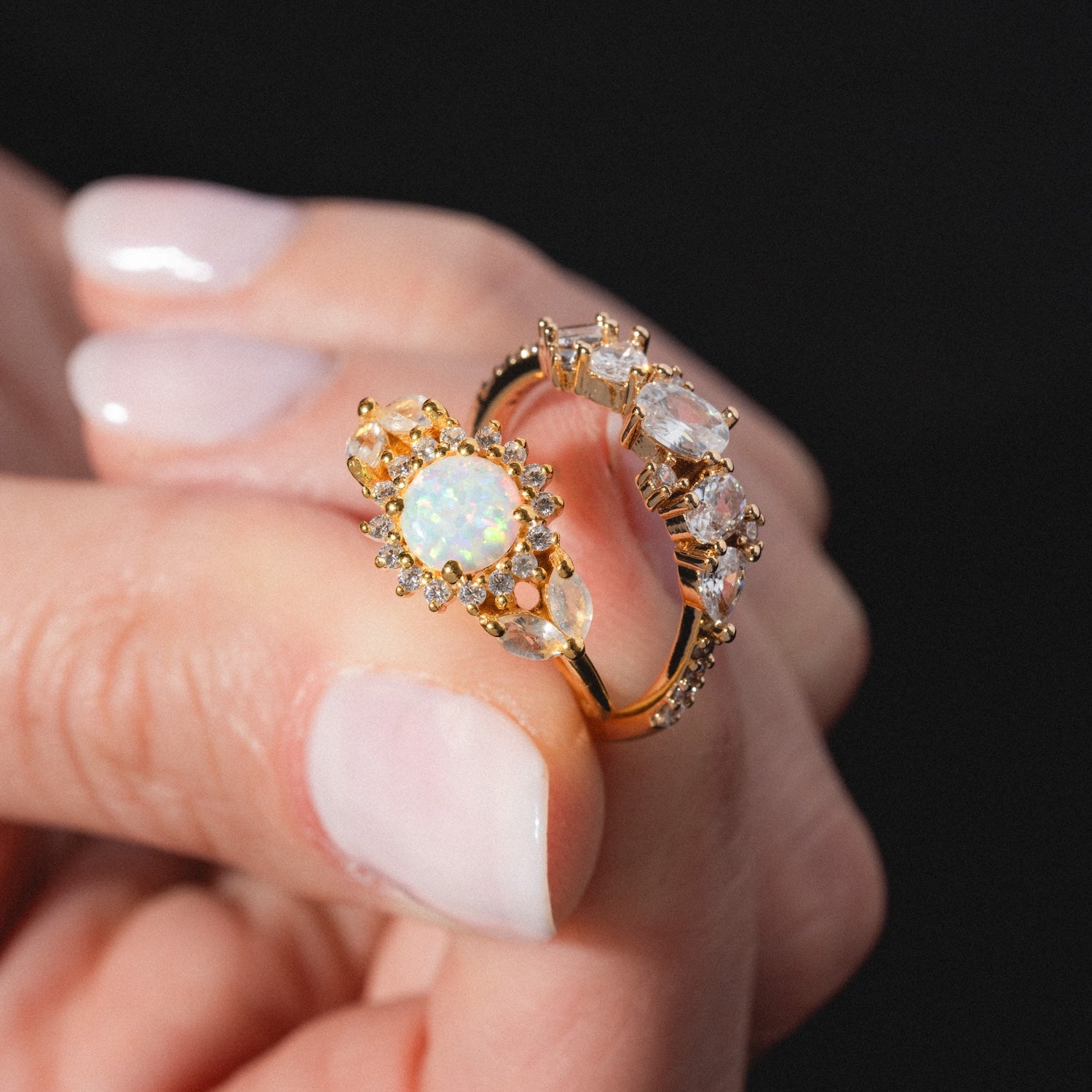 A hand with manicured nails holds the Statement Crystal Cluster Ring, featuring a gold band with a sparkling opal center, surrounded by diamonds and clear gemstones. The background is dark and out of focus.