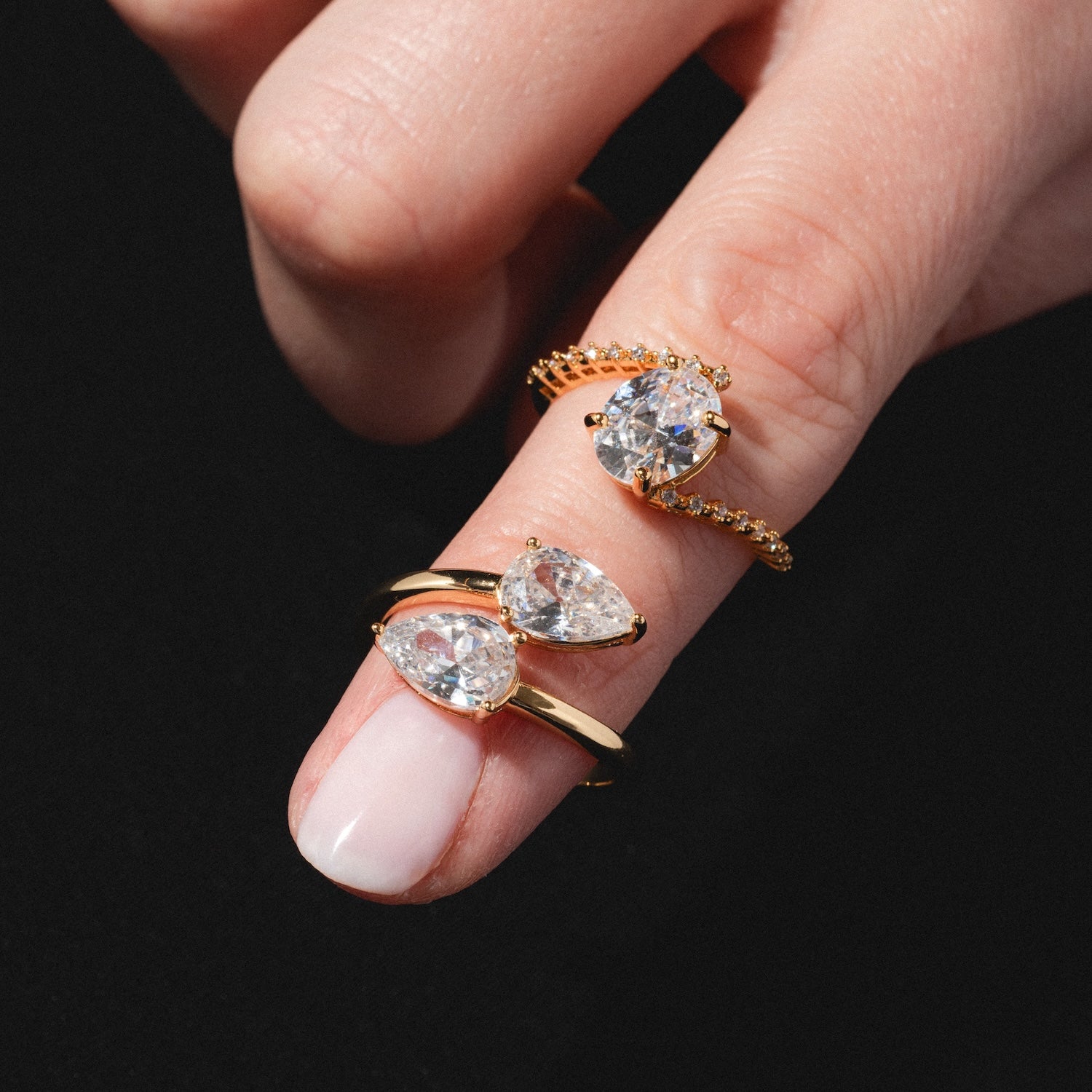 A close-up of a hand wearing three Tilted CZ Rings, each featuring large, clear oval and pear-shaped gemstones on gold bands, set against a black background.